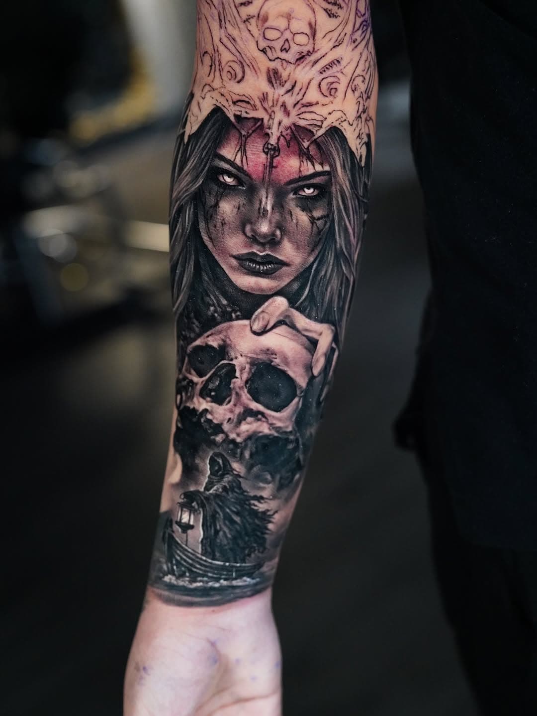 Dark realism forearm tattoo featuring a woman's face with dramatic eyes, skull below, and intricate gothic patterns above in black and grey