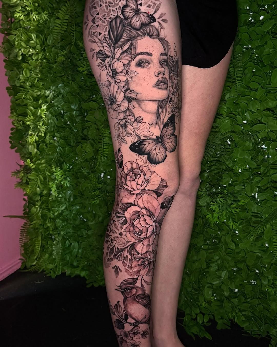 Black and grey leg tattoo featuring a woman's portrait surrounded by flowing flowers, butterflies, and a bird in illustrative style