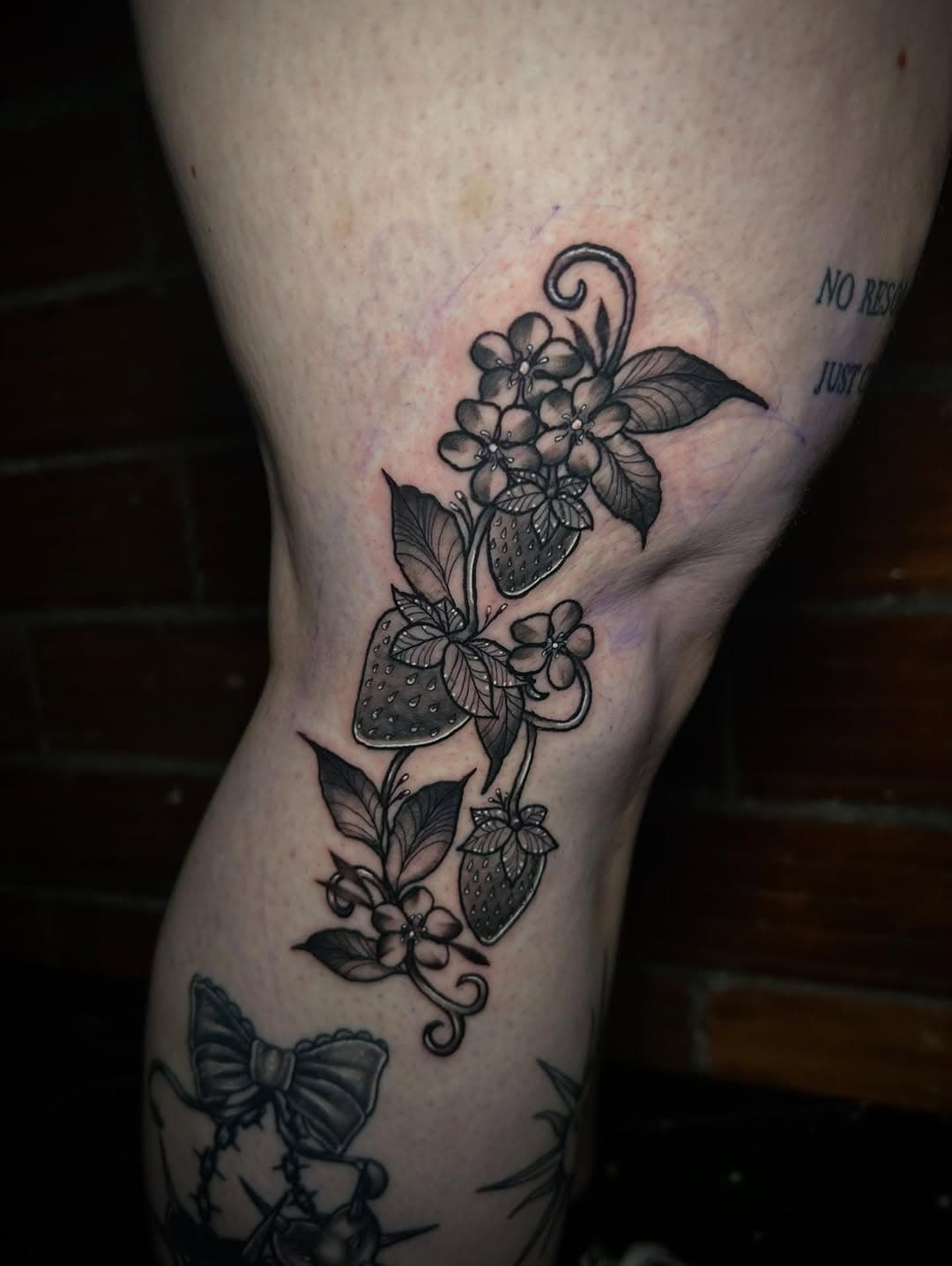 Black and grey illustrative tattoo of strawberry plant with flowers, leaves, and fruit in decorative botanical style on leg