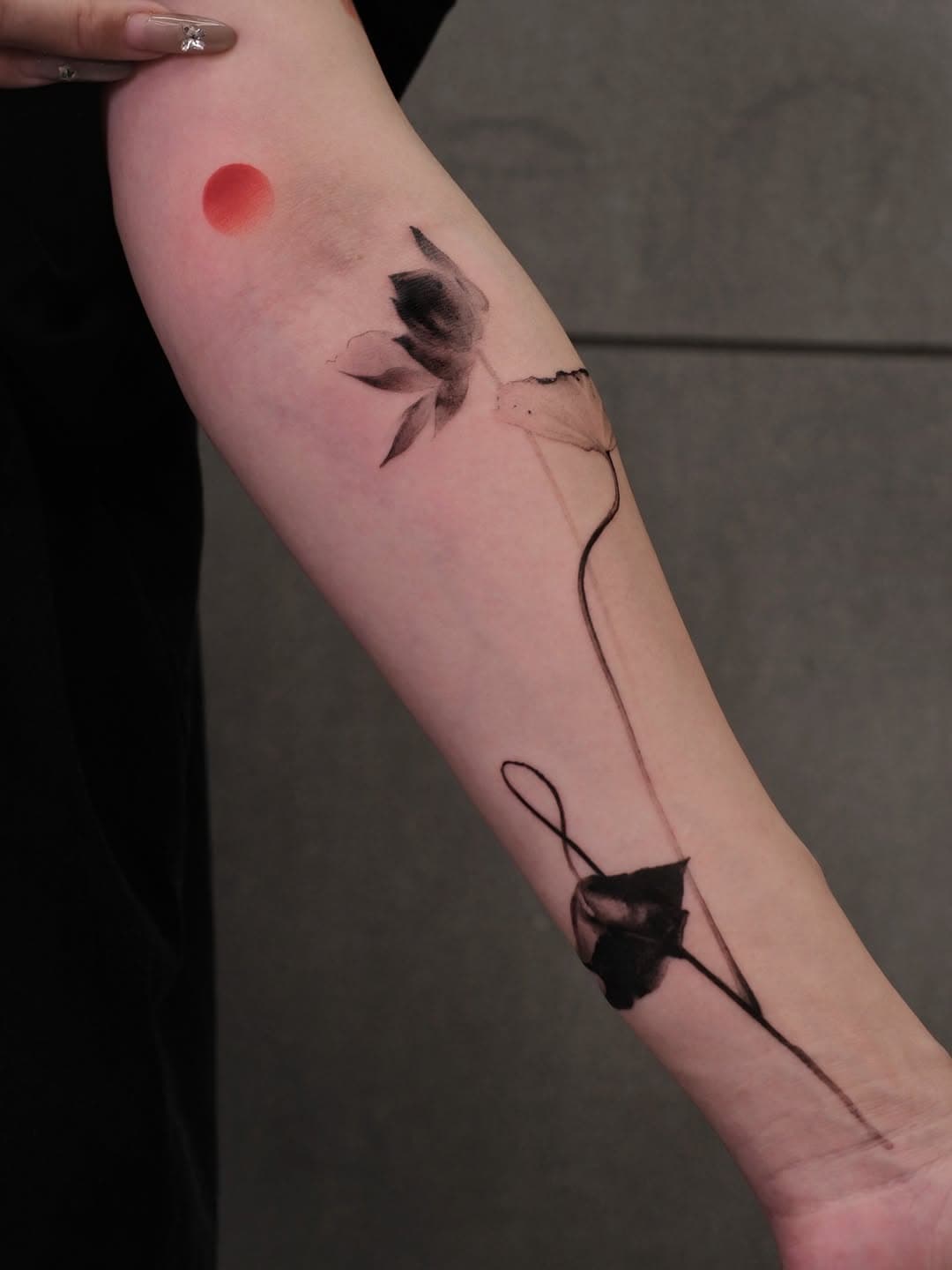 Minimalist blackwork tattoo of a rose stem with leaves and a red dot sun on forearm in fine-line illustrative style