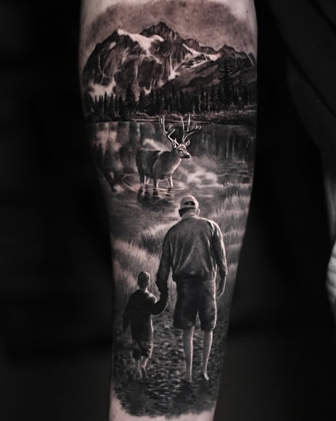Black and grey realism tattoo depicting man and child walking toward deer in mountain lake landscape with snow-capped peaks