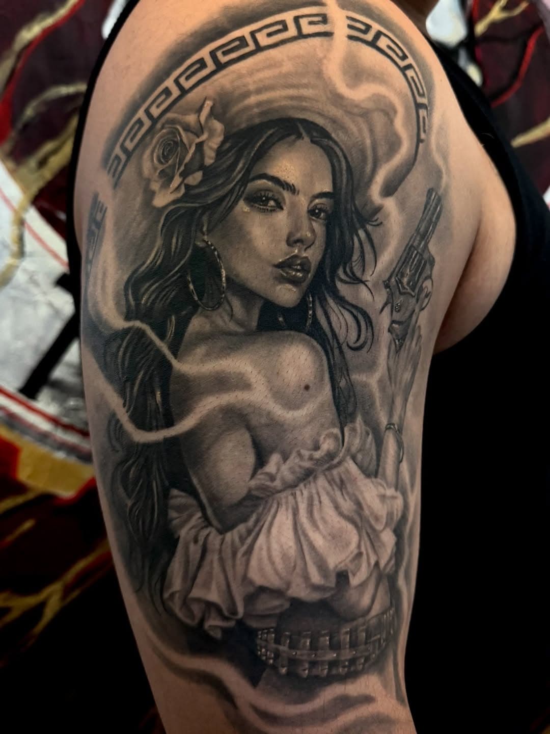 Black and grey realism tattoo of a woman with long flowing hair, rose detail, Greek key pattern halo, and pyramid element on upper arm
