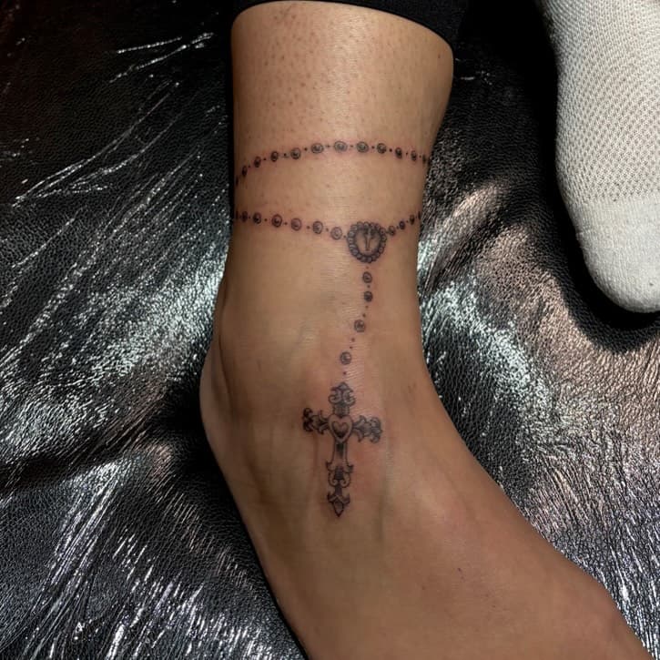 Black and grey rosary beads tattoo wrapping around ankle with ornate cross pendant dangling down foot in neo-traditional style